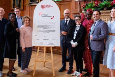 The Sanctuary Team: from L to R, John O’Neill, Tiwalade Olatunbosun, Raymond Miller, Sandra Griffiths, Maurice Macartney, Federica Ferrieri, Israel Eguaogie, Mehrshad Esfandiari, Aileen Cummins standing either side a large white cathedral window shaped board, with certification of University of Sanctuary status.  Staff are wearing an array of suits, dresses, national dress and smart office wear.